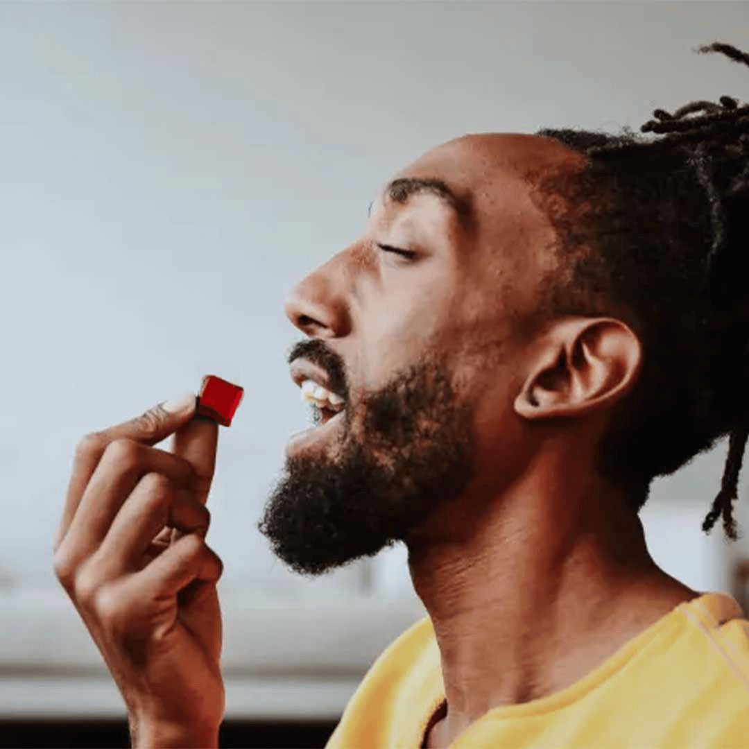 A man tilts his head back and eats a small gummy supplement, holding it near his mouth against a neutral background.