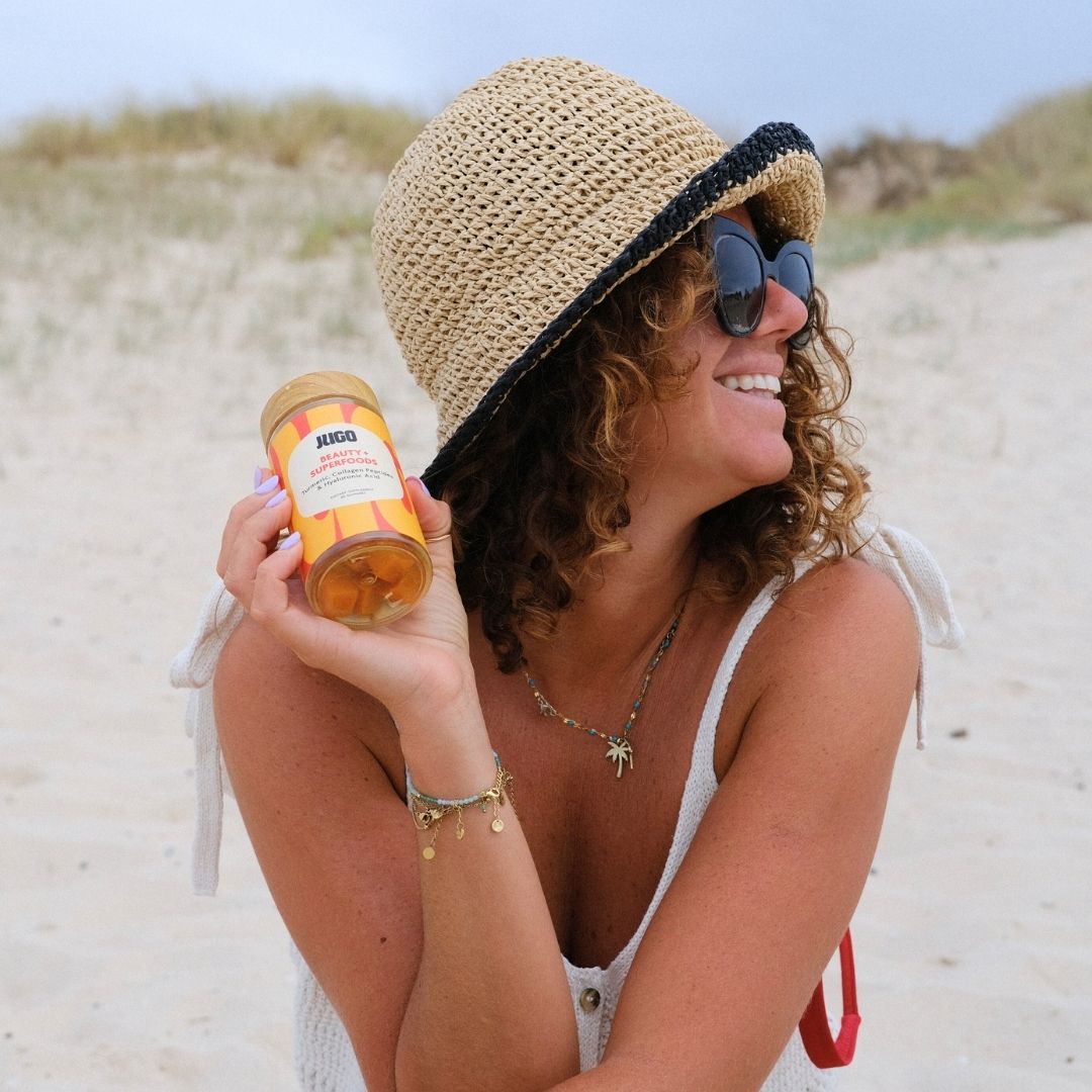 A woman at the beach smiles while holding a Jugo Superfoods gummy bottle, wearing sunglasses and a straw hat.