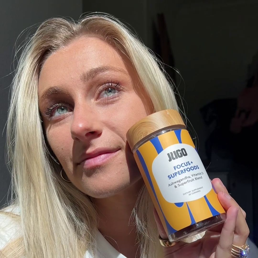 Close-up lifestyle photo of a woman holding a Jugo Focus Superfoods gummies bottle near her face in soft indoor lighting.