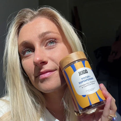 Close-up lifestyle photo of a woman holding a Jugo Focus Superfoods gummies bottle near her face in soft indoor lighting.