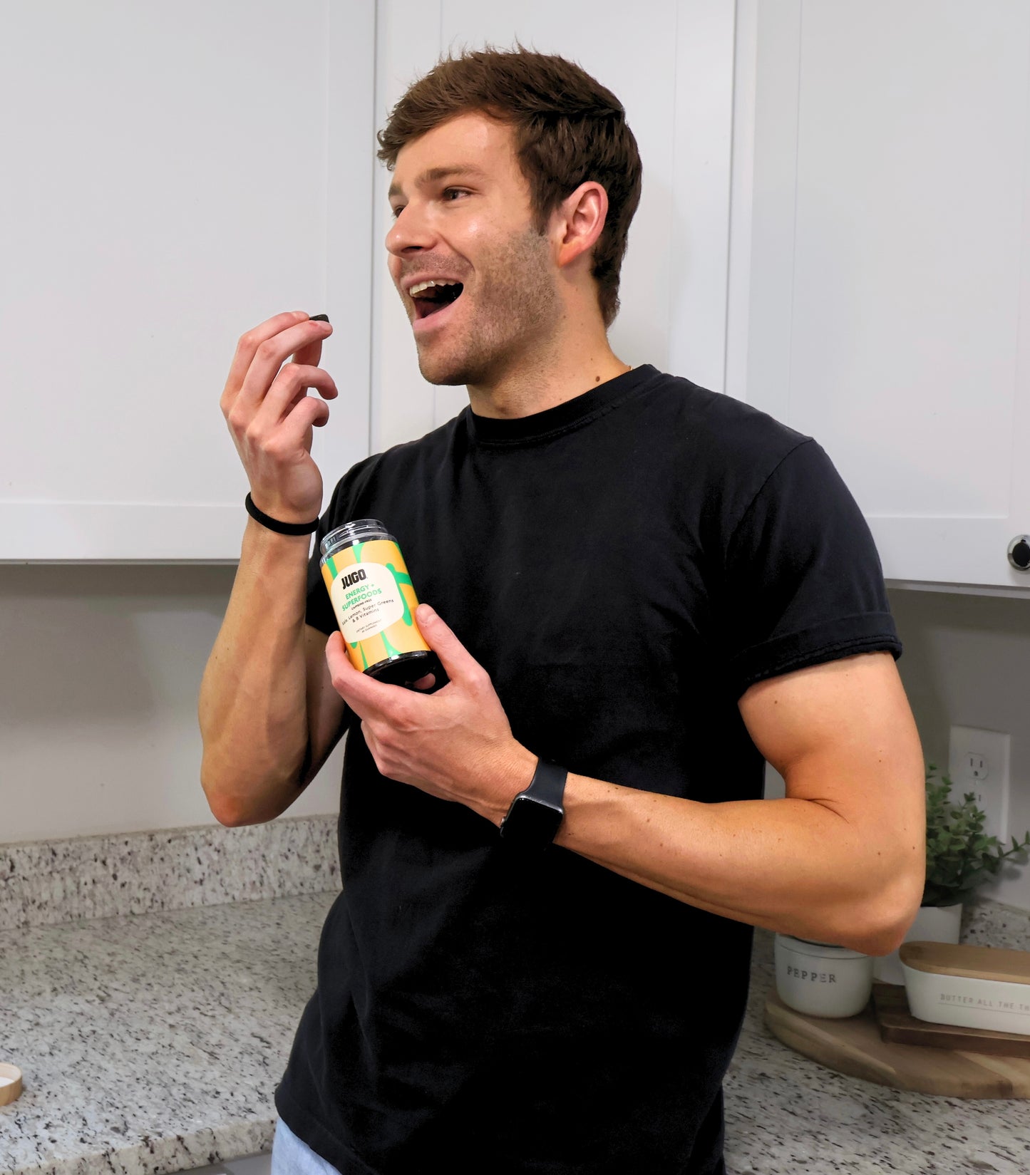 A man indoors smiles while eating a gummy and holding a Jugo Energy Superfoods bottle.