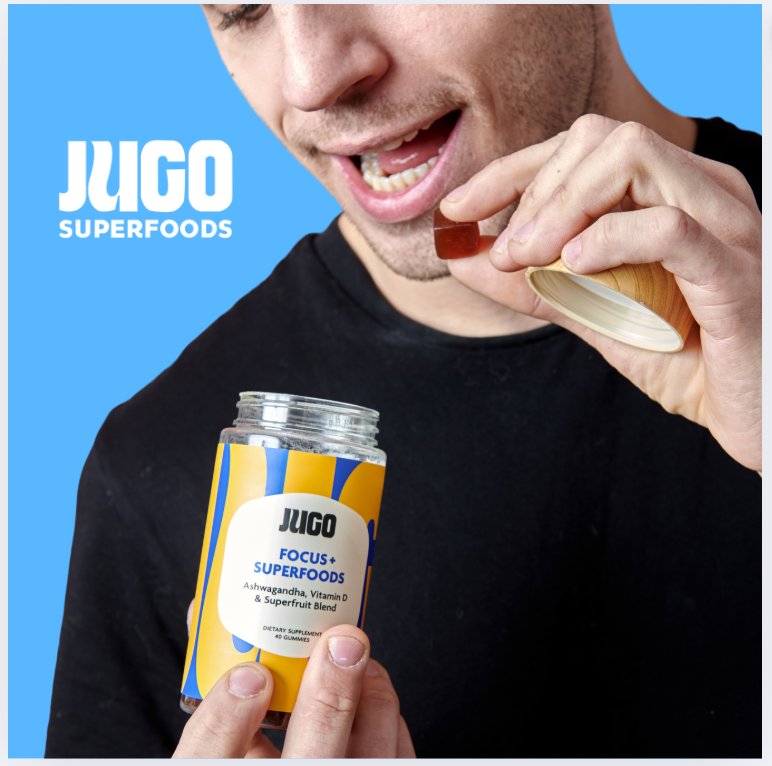 A man smiles while eating a gummy and holding a Jugo Focus Superfoods bottle against a bright blue background.