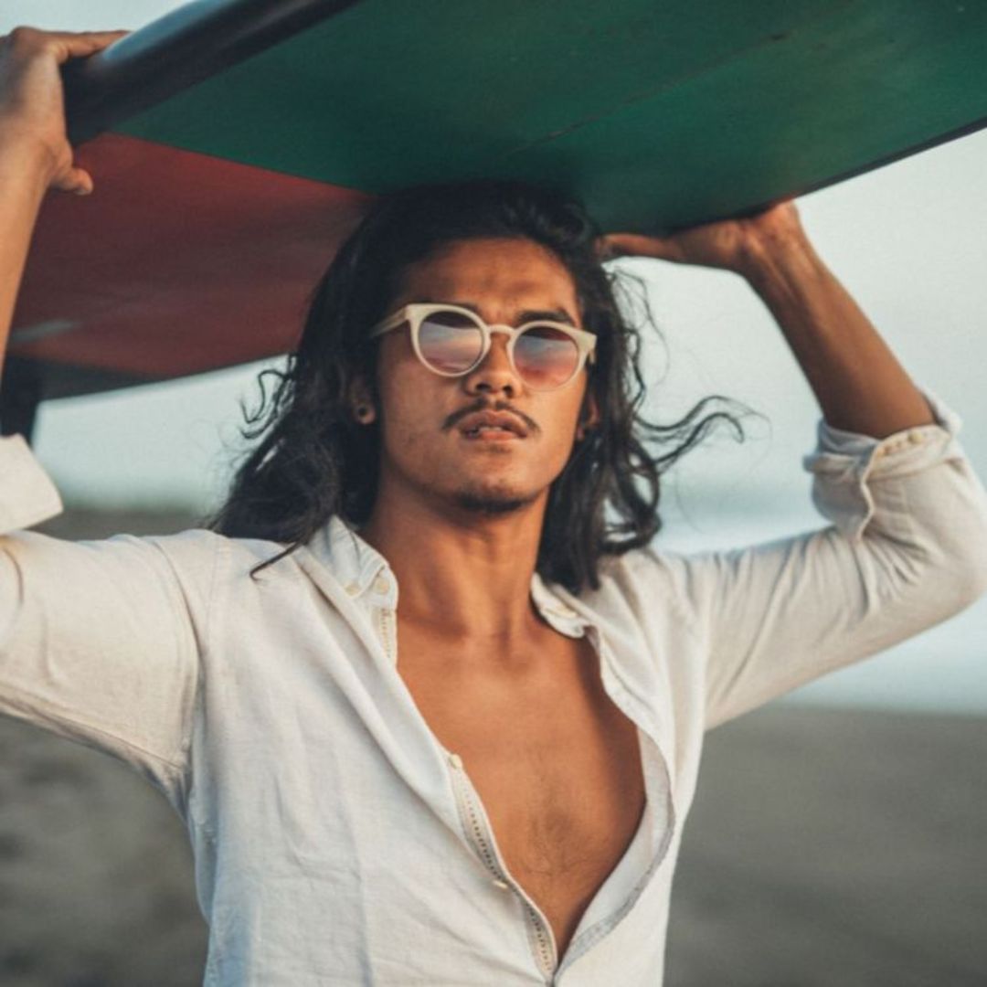 Lifestyle image of a person holding a surfboard overhead at the beach, standing in shallow water under a bright sky.
