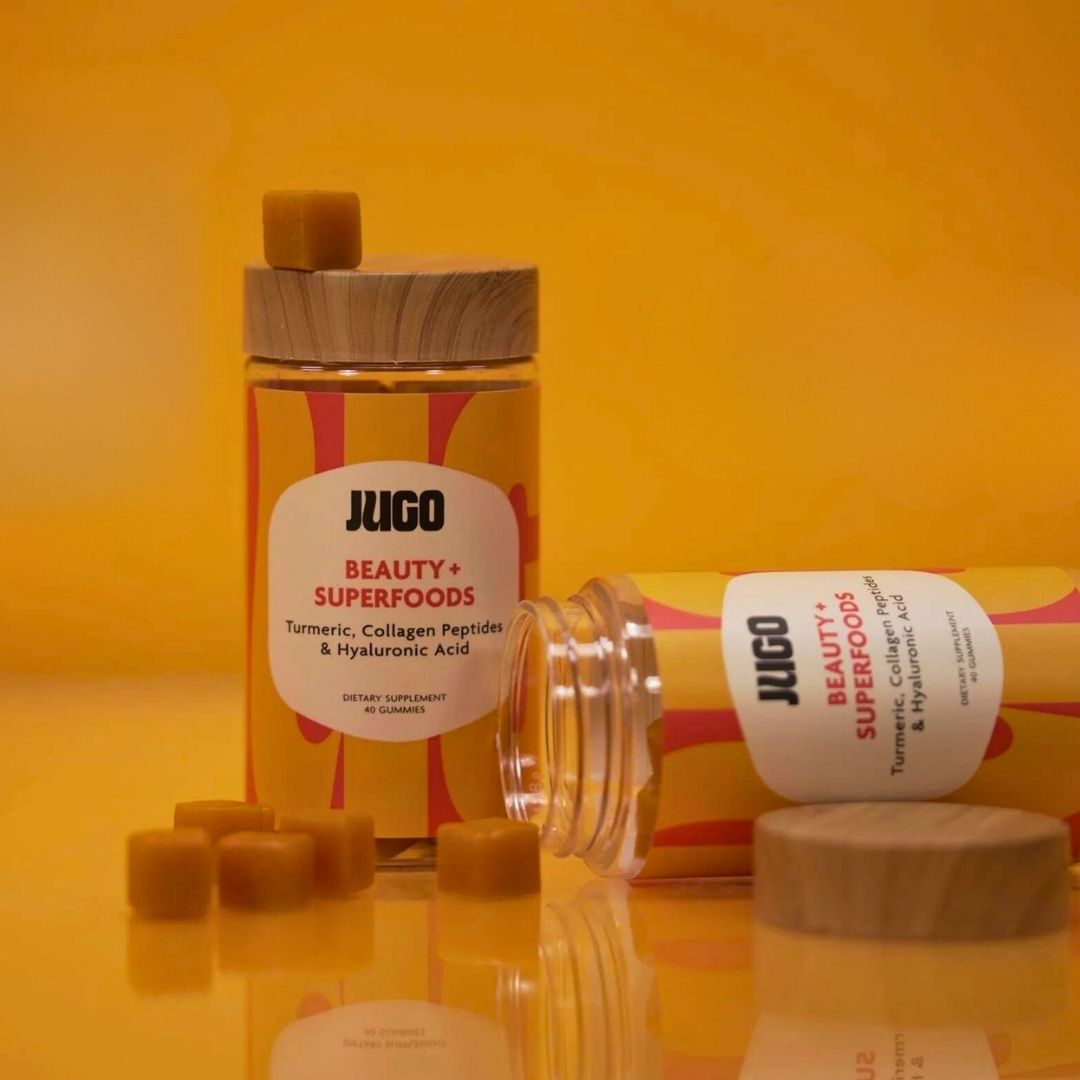 Studio product shot of a Jugo Beauty Superfoods bottle surrounded by stacked gummies on a warm orange background.