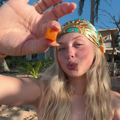 A woman outdoors holds a gummy up to the camera while puckering her lips, with blue sky and sunlight in the background.
