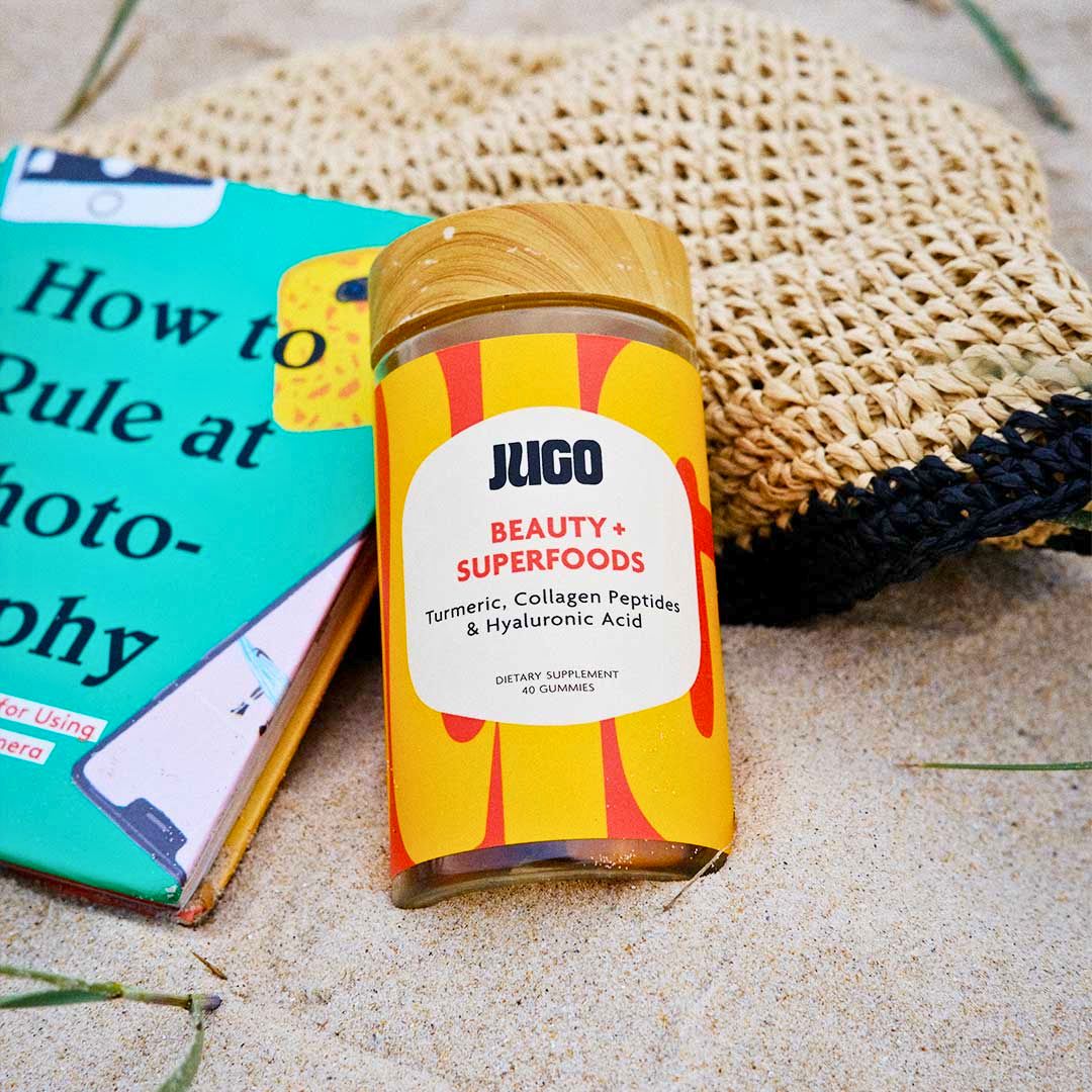 A jar of Jugo Beauty Superfoods gummies styled on a woven bag next to a book on a sandy surface in a lifestyle setting.