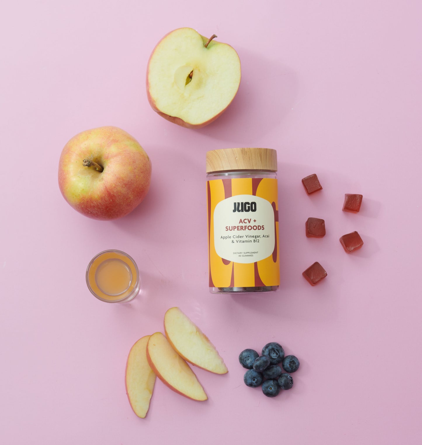 Flat lay of a Jugo ACV Superfoods bottle surrounded by apples, blueberries, apple slices, and gummies on a pastel pink background.