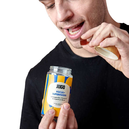 A man smiles while eating a gummy and holding a Jugo Focus Superfoods bottle against a bright blue background.